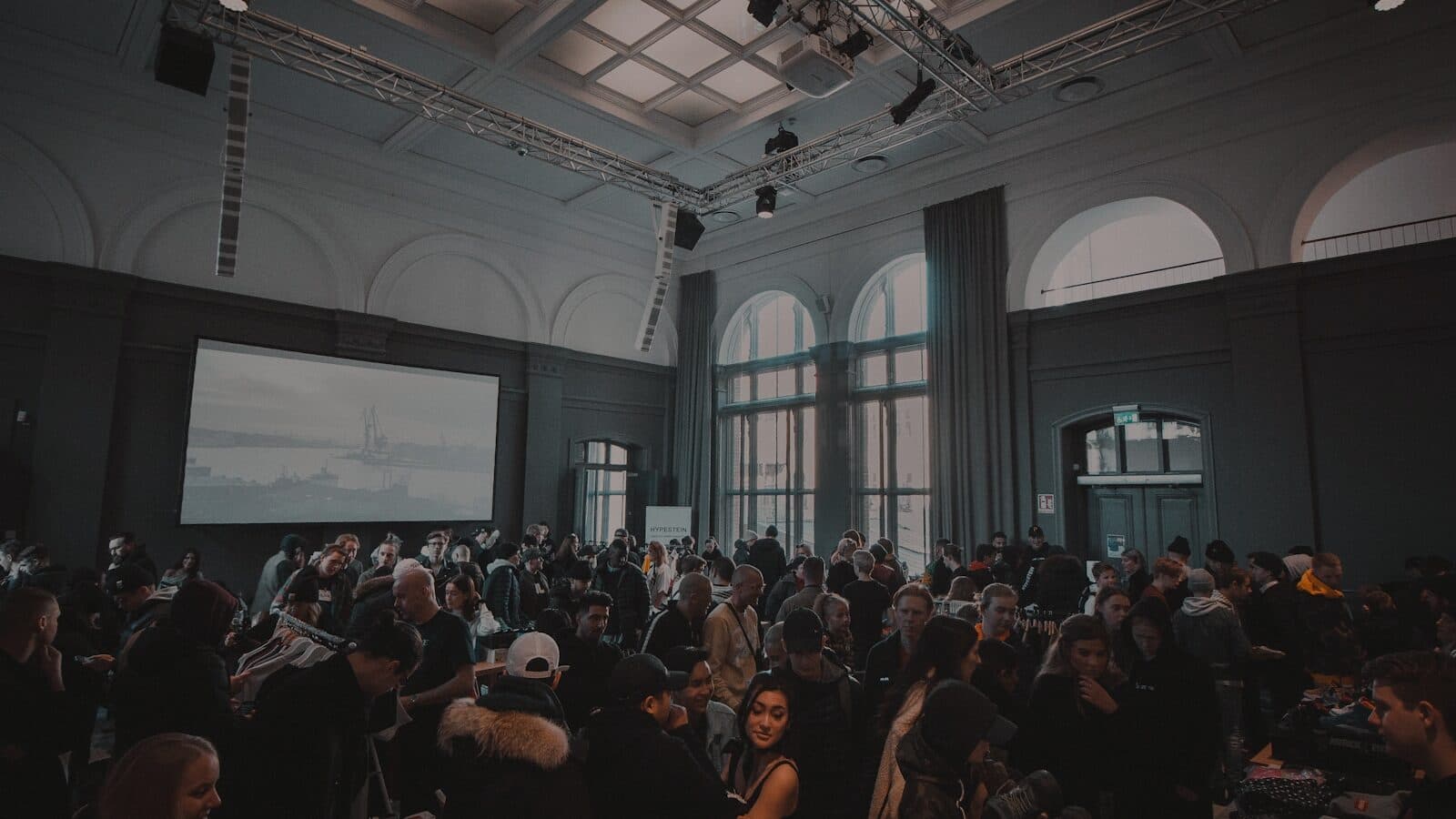 A packed event hall under lighting truss, a projection screen at one end and the floor full of people.