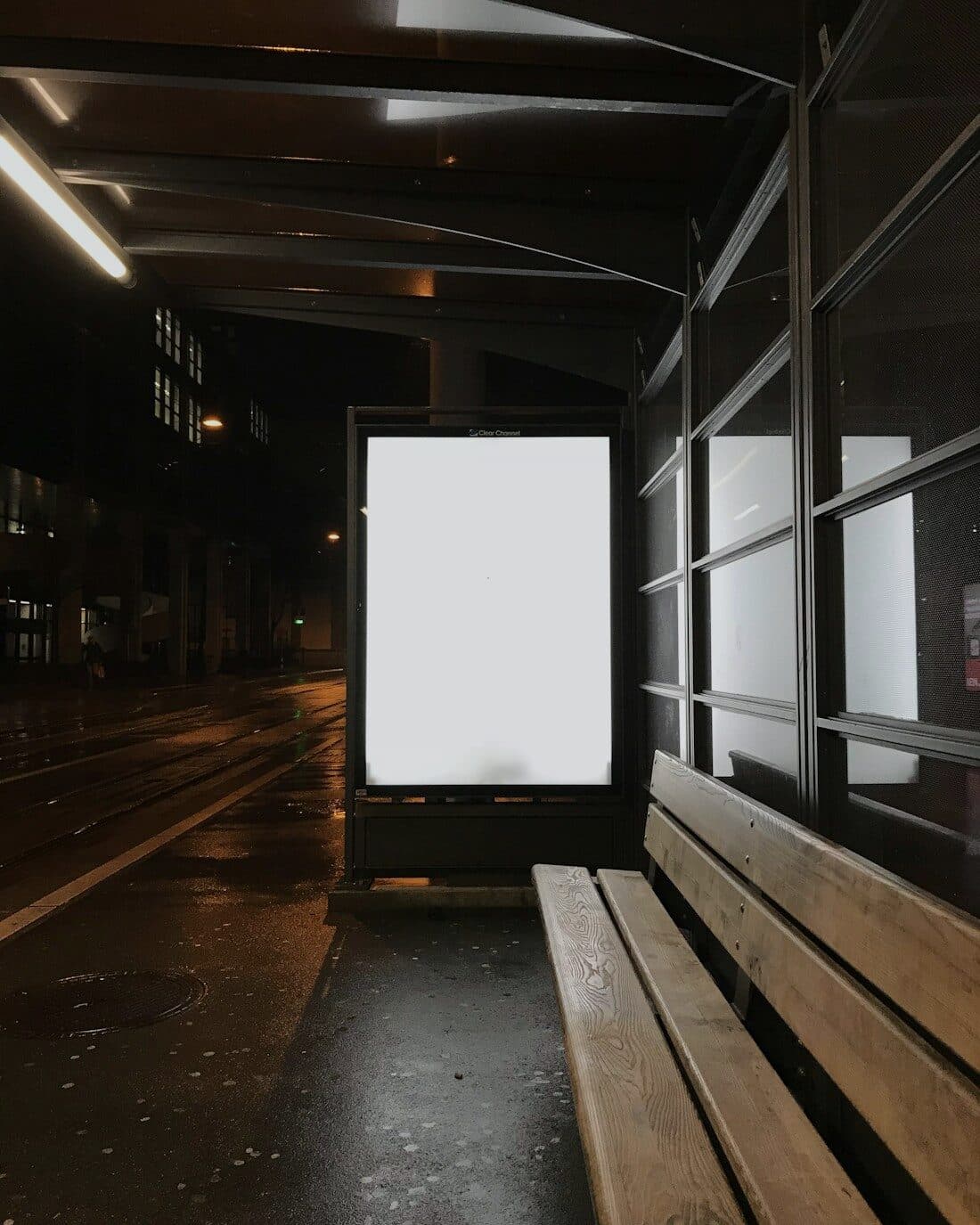 A blank lit advertising panel standing at a bus stop on a wet street at night
