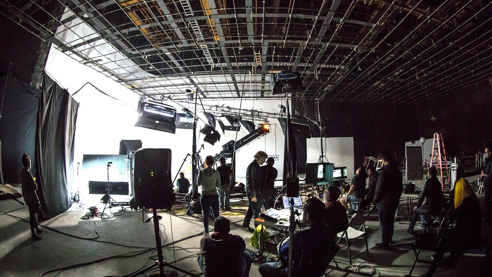 A crew at work on a lit soundstage, lighting rigs and a camera crane up around a white backing